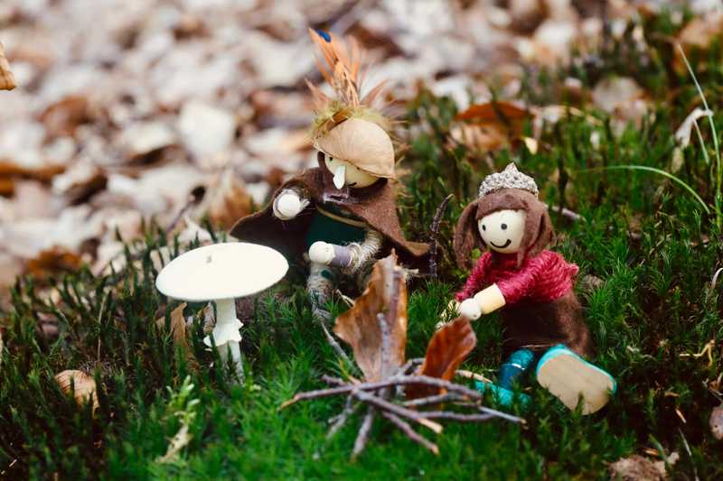 Zwei kleine Filzfiguren sitzen an einem Mini-Lagerfeuer neben einem weißen Pilz im Moos