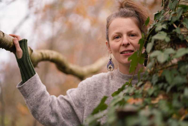 Mandy Falke im herbstlichen Wald, lächelnd, die Hand auf einem Ast
