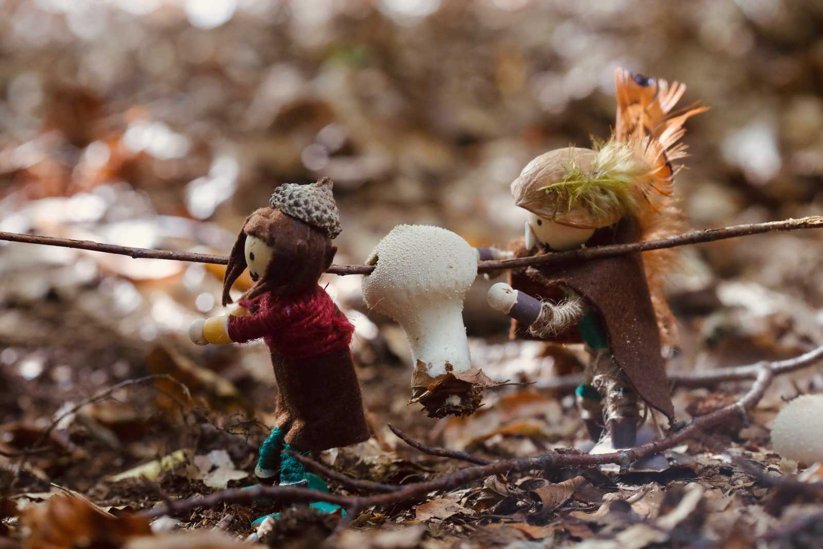 Zwei kleine Filzfiguren tragen einen weißen Pilz an einer Stange durch das Herbstlaub
